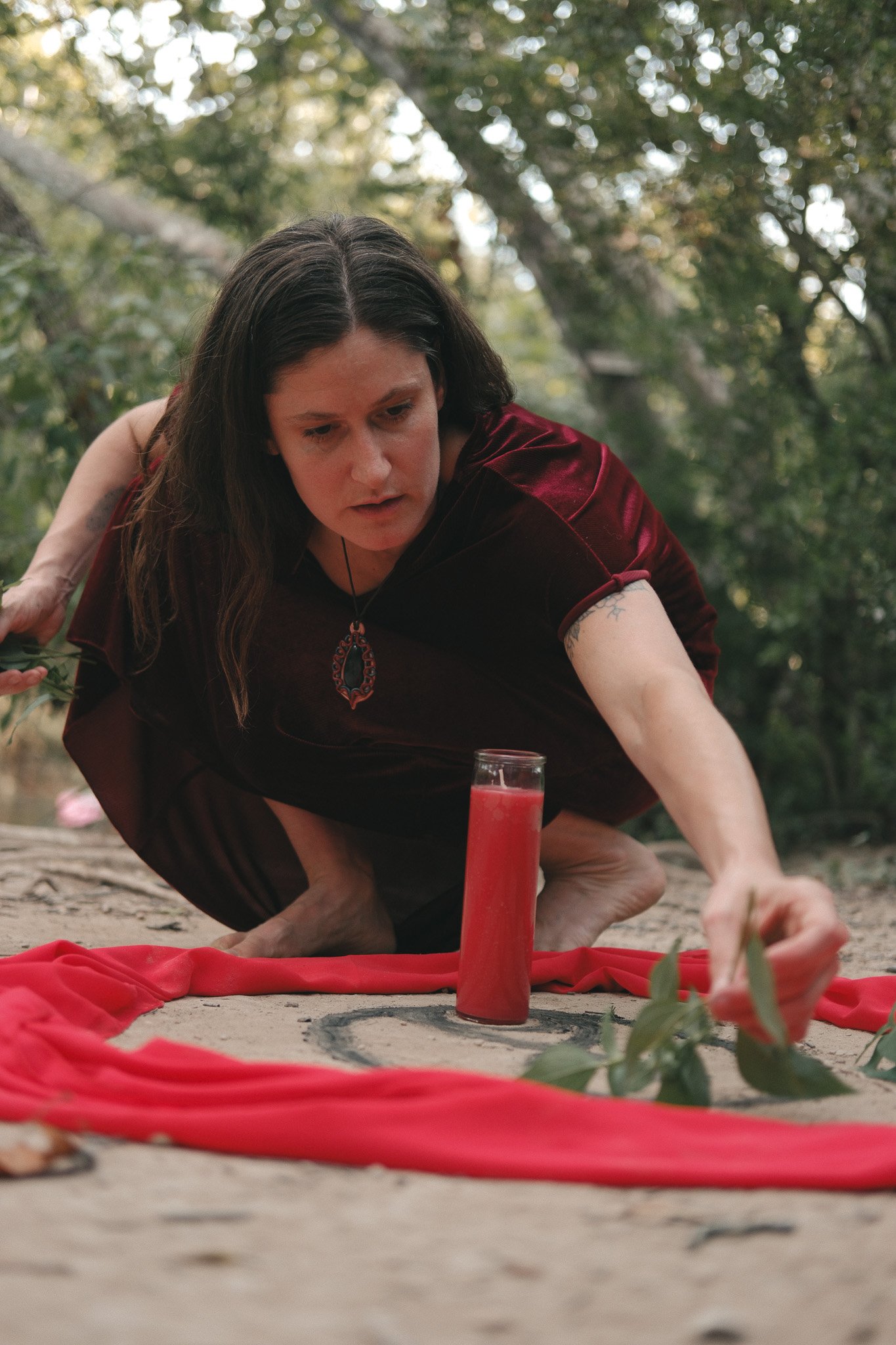 A woman dressed in a dark red velvet garment is positioned on her hands and knees outdoors on a sandy surface, reaching towards a red candle holder on a red fabric cloth draped on the ground. The background is filled with green trees and daylight filtering through the foliage.