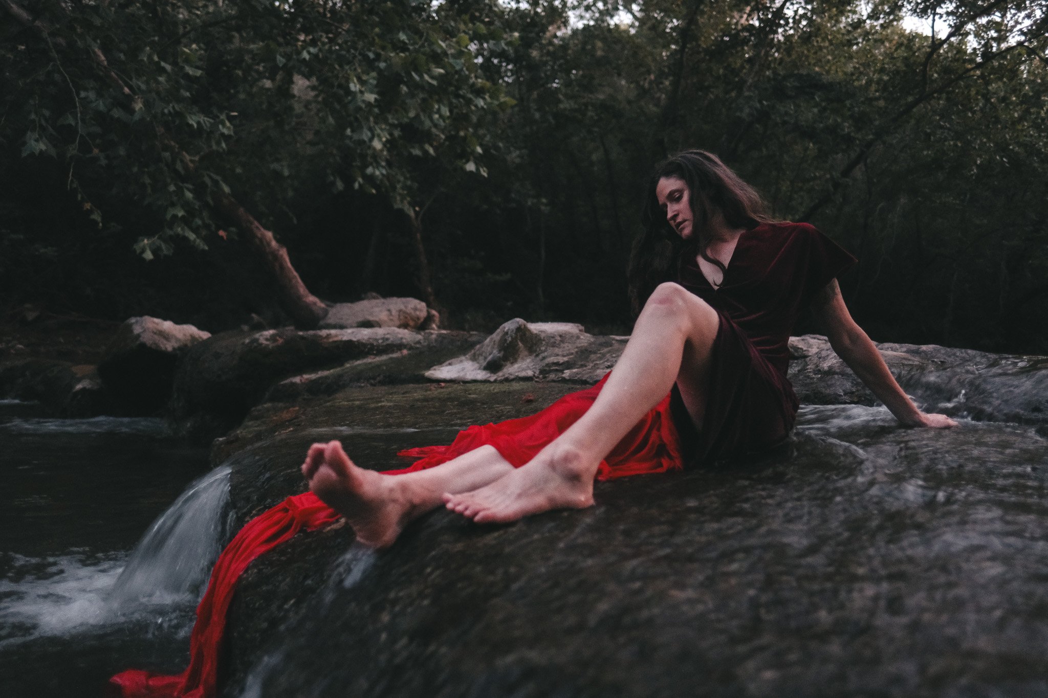 A woman in a dark velvet dress sitting on rocks by a stream surrounded by trees. A bright red fabric is draped over her leg.