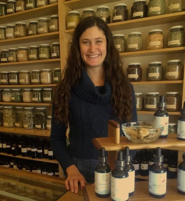 A woman with long curly brown hair smiling behind a wooden counter, in front of a shelf filled with glass jars of herbs, in an apothecary setting.