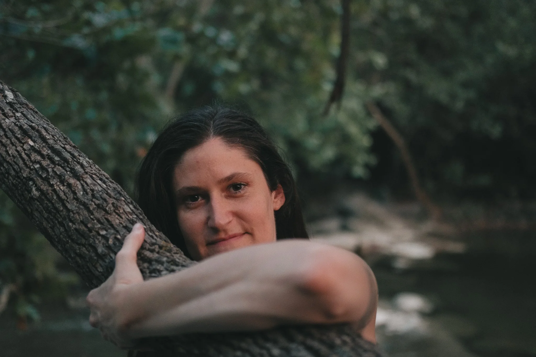 A woman with dark hair and fair skin partially hidden behind a tree branch in a forest, with green leaves and trees in the background.