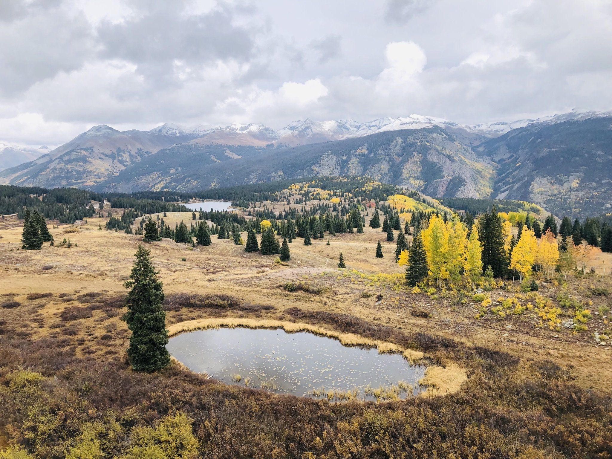 Scenic view of snow-capped mountains with a cloudy sky, a forested hill with changing autumn-colored trees, and a small pond in the foreground.