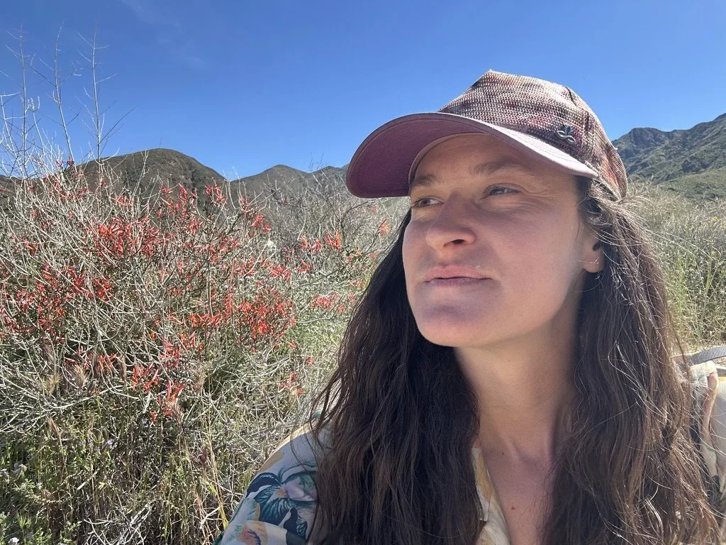 Woman with long dark hair wearing a pink cap outdoors in a mountainous area with dried bushes and a blue sky.