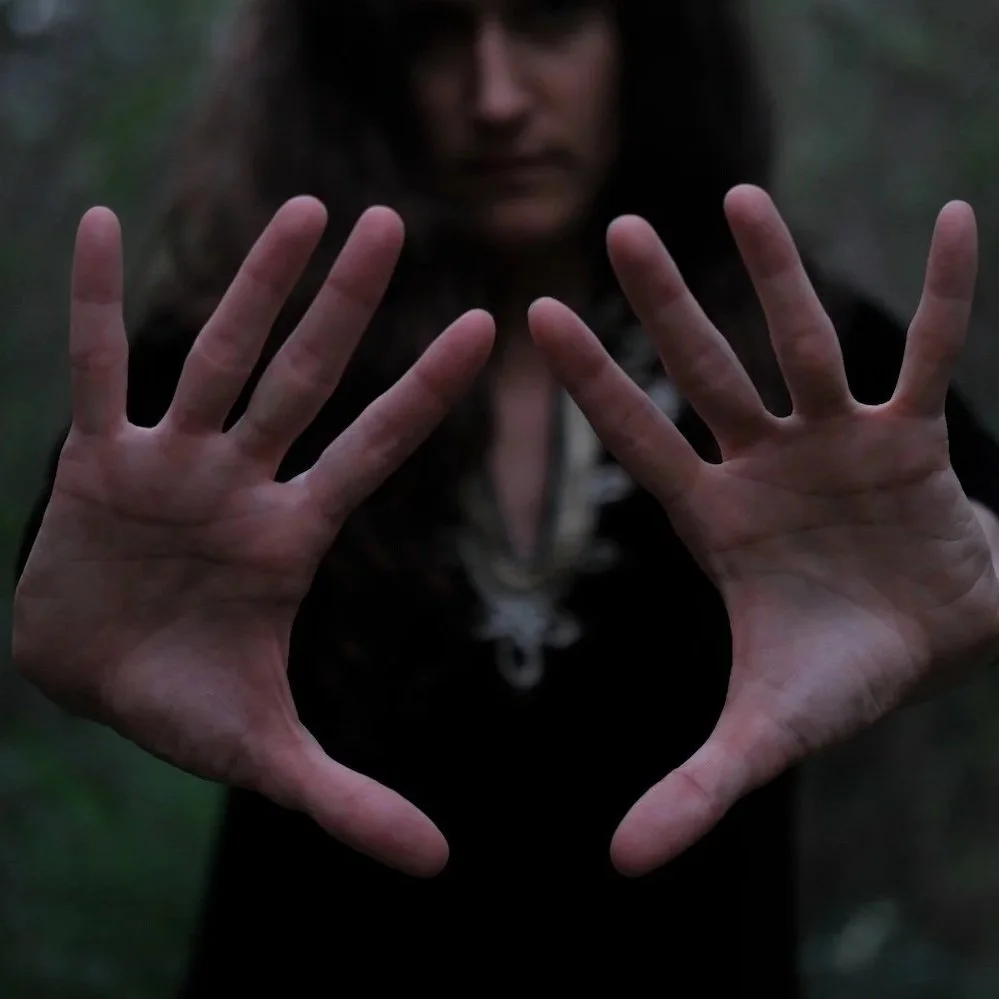 A person with long dark hair holding both hands up towards the camera. The background is blurred and outdoors.