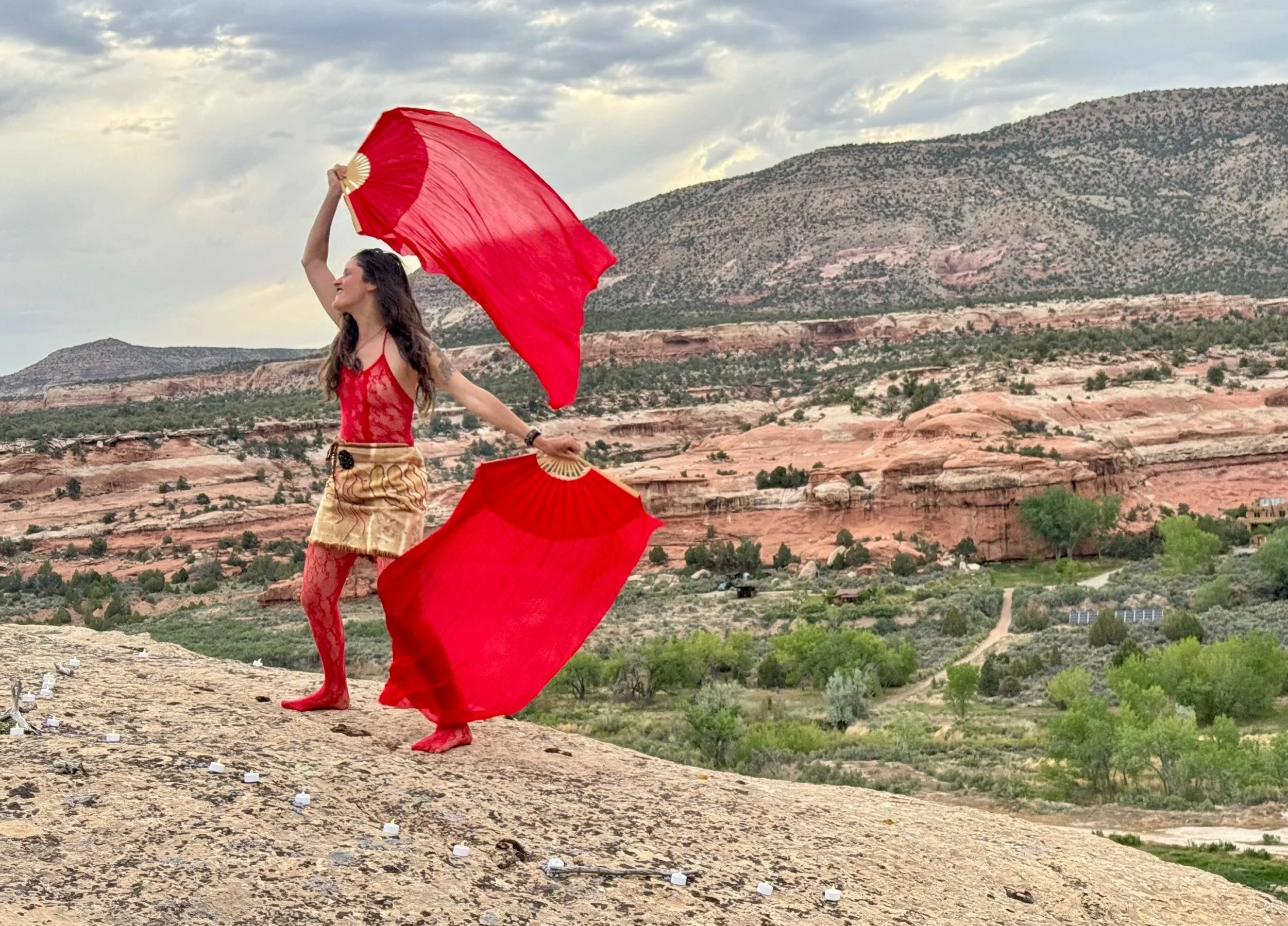 A woman in red lace top and beige skirt holds red silks while dancing on a hill with a desert landscape and mountains in the background.