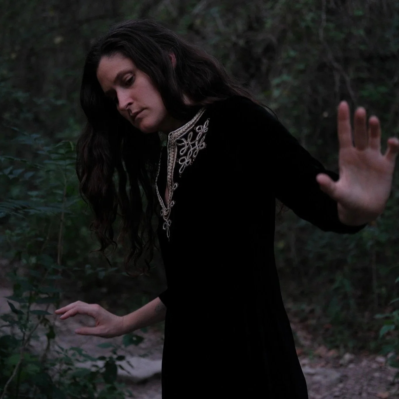 Woman with long dark hair and a black dress with intricate embroidery, standing in a wooded area, with her hands extended as if sensing or feeling her surroundings.