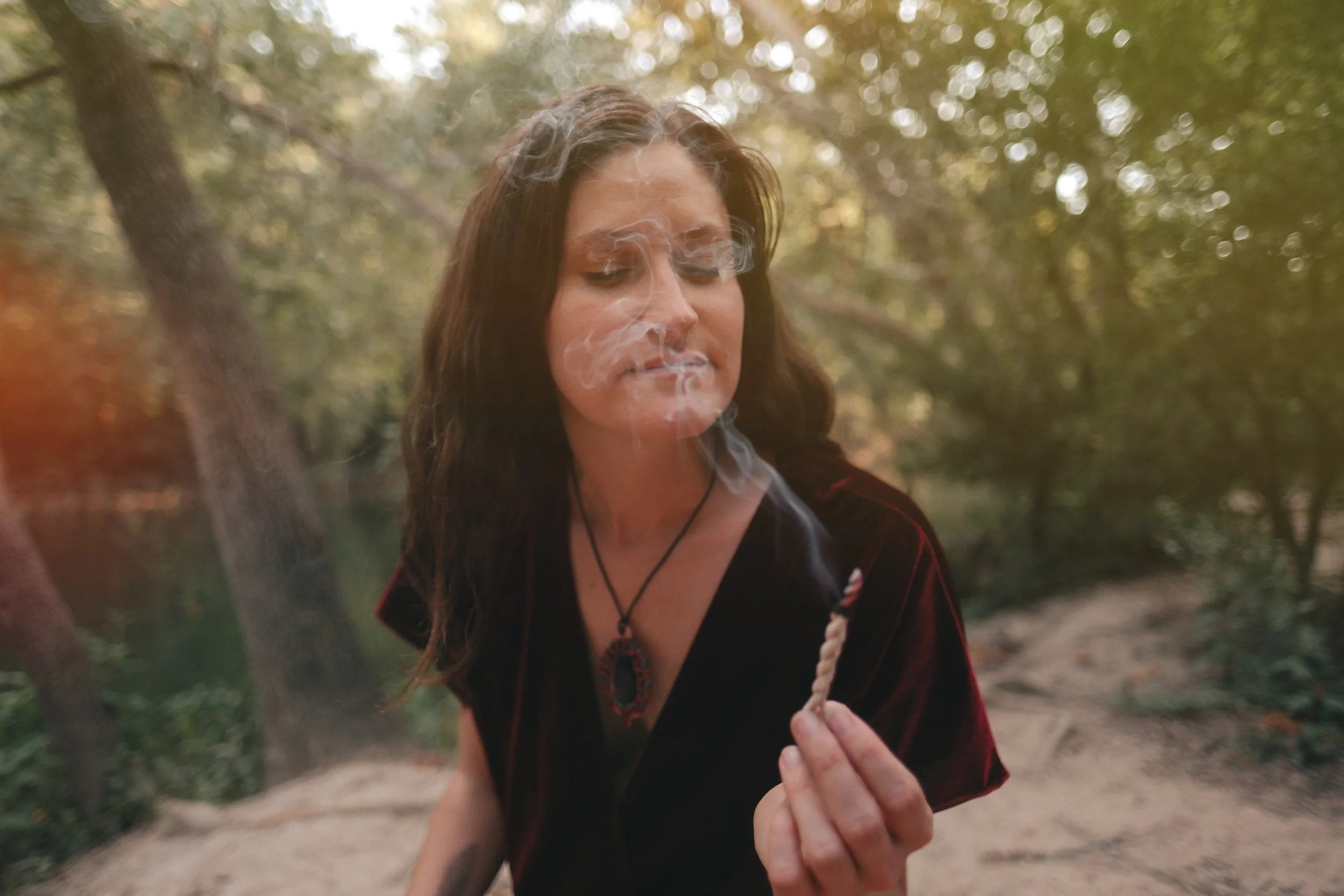 Woman with long hair in a forest, holding a lit coil of cedar incense. The smoke wafts in her face.