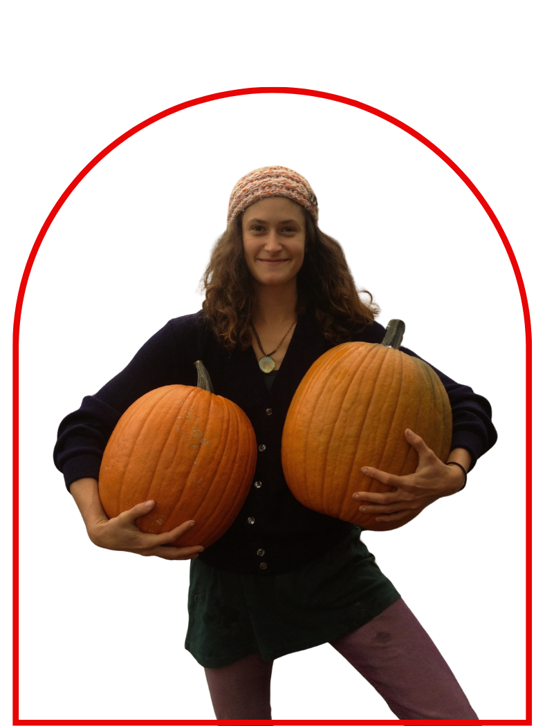 Young woman smiling, wearing a knit hat and dark clothing, holding two large pumpkins.