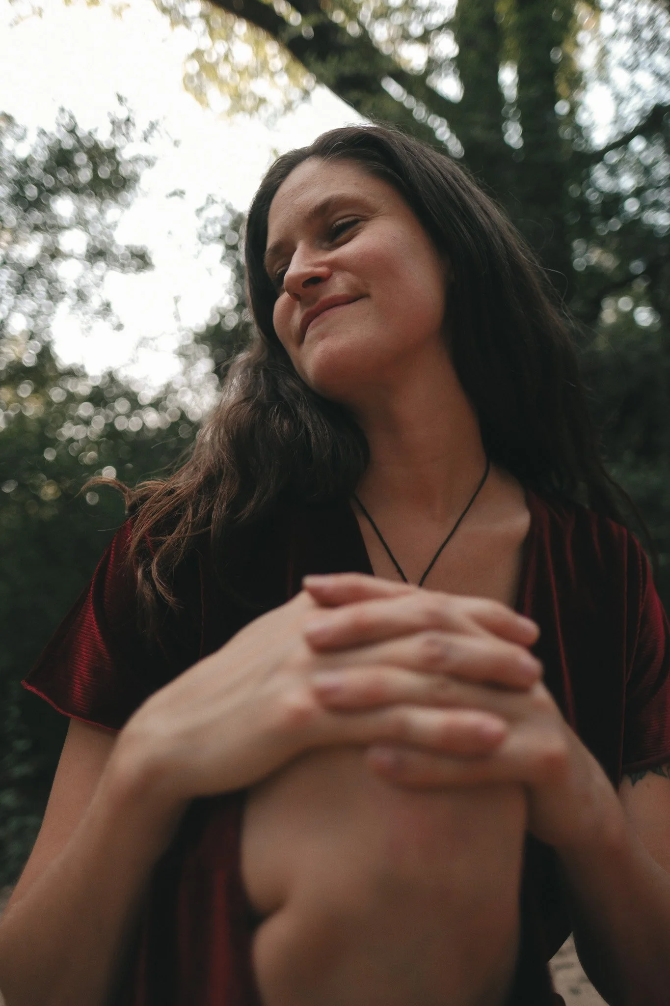 A woman with long dark hair smiling with her eyes closed outdoors, wearing a dark dress with red details and a thin black necklace, with blurred trees and sky in the background.