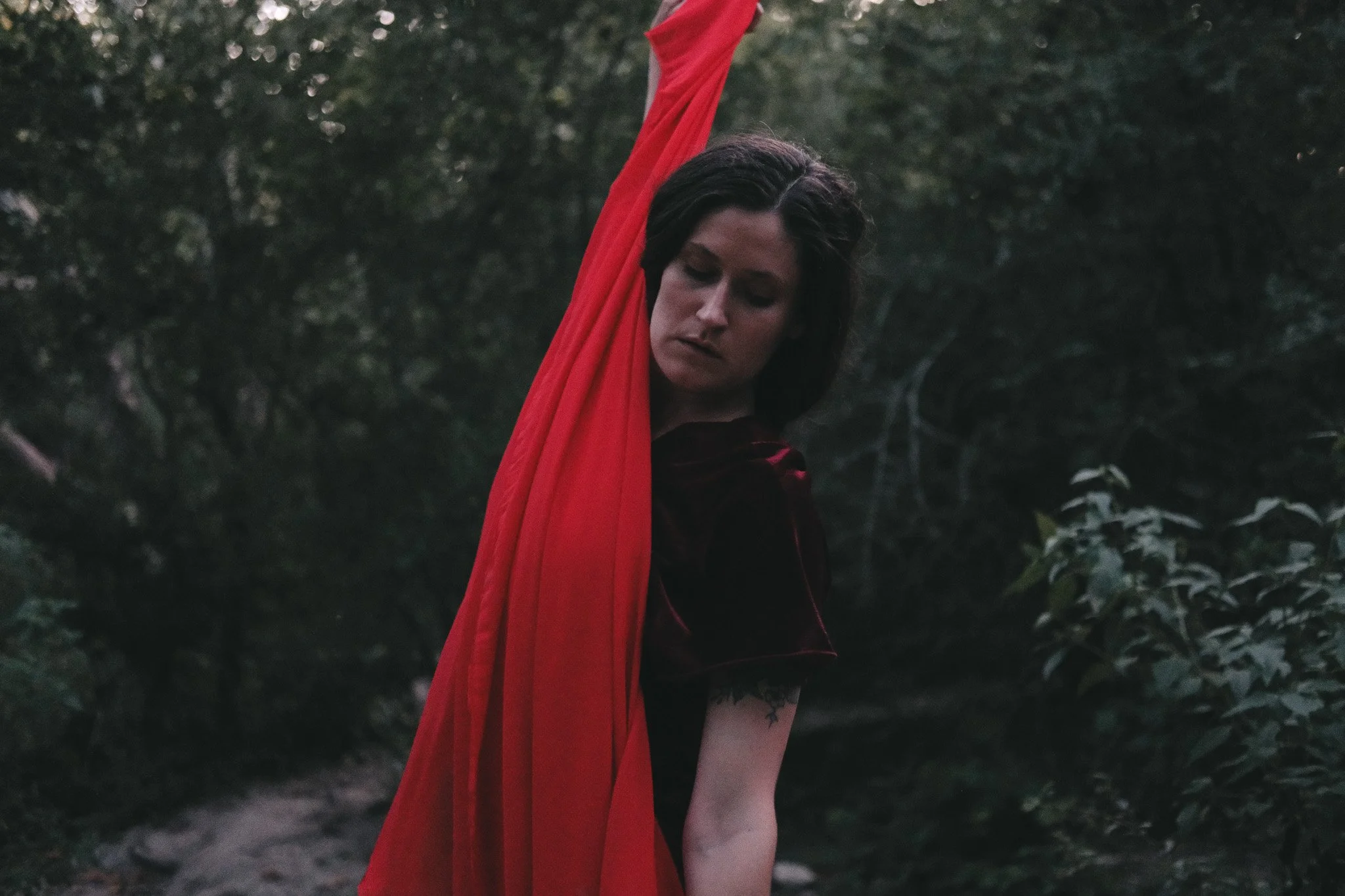 A woman with dark hair and pale skin standing outdoors in a natural setting with trees and bushes, wearing a dark dress outfit and stretching a big piece of red fabric.