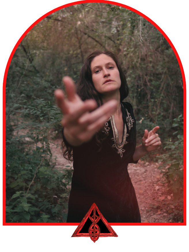 A woman with long dark hair in a black embroidered dress standing in a wooded area, reaching out with her right hand towards the camera.