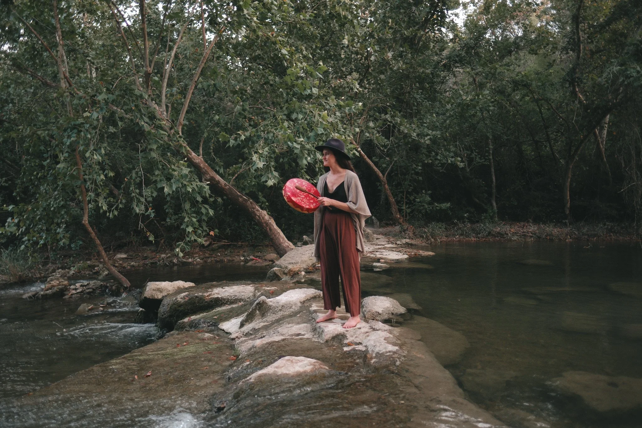 A woman standing on rocks by a river playing a red drum, with dense green trees in the background.