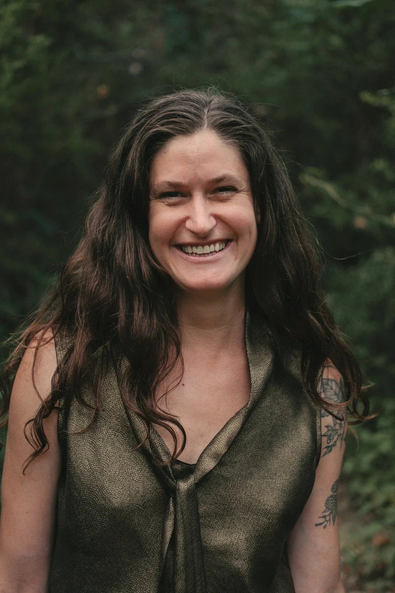 A woman with long, wavy dark hair smiling outdoors with a green leafy background.