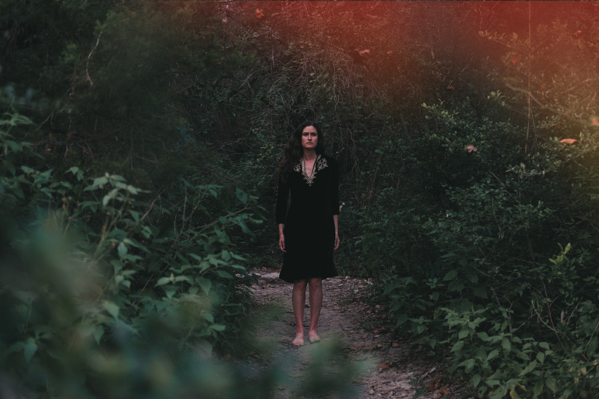 A woman with long dark hair in a black dress standing barefoot on a dirt path surrounded by dense green foliage in a forest setting.