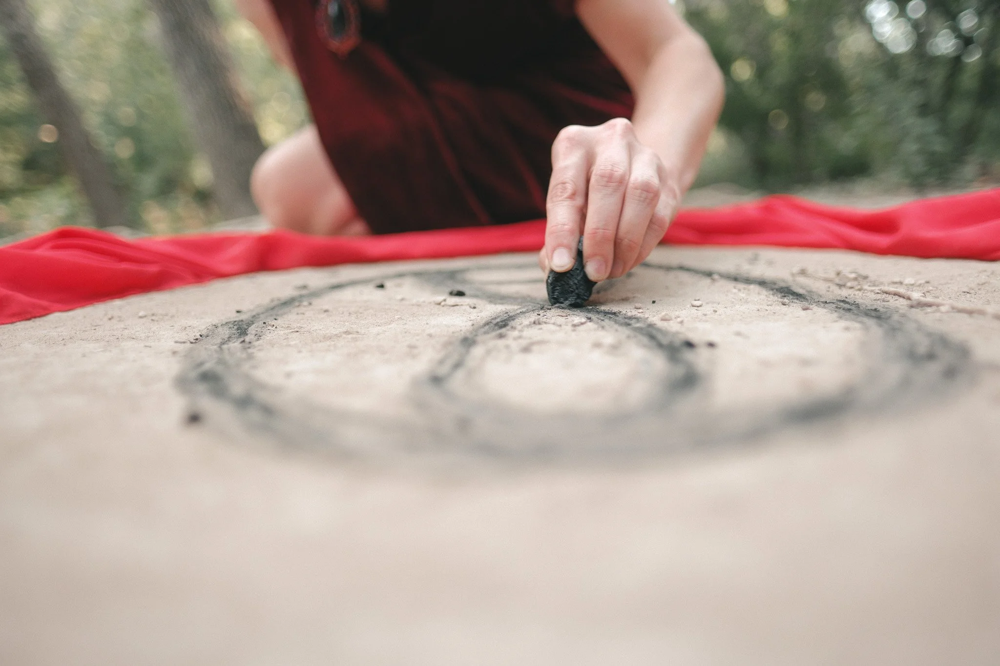 A person drawing a sigil on the ground outdoors with a piece of charcoal, surrounded by a red cloth.