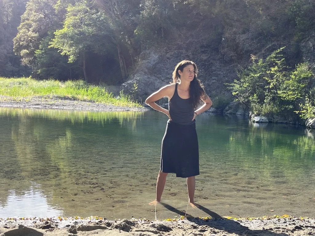 A woman standing in shallow water near a forested riverbank, wearing a tank top and skirt with hands on her hips, gazing into the distance.
