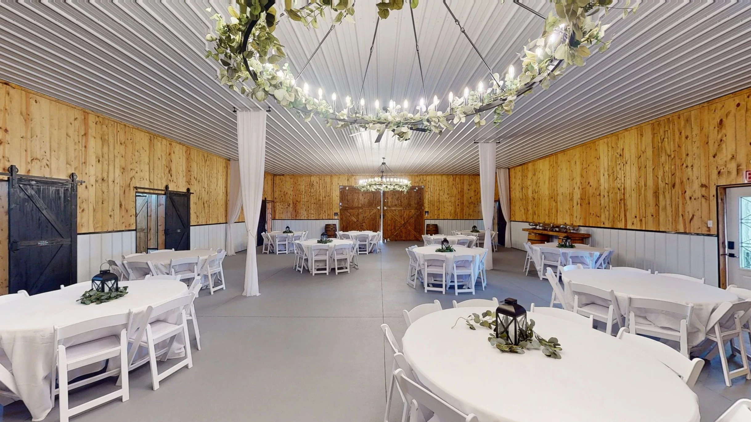 Elegant event hall with round tables covered in white tablecloths, decorated with greenery and lantern centerpieces, wooden walls, white chairs, and a floral chandelier hanging from the ceiling.