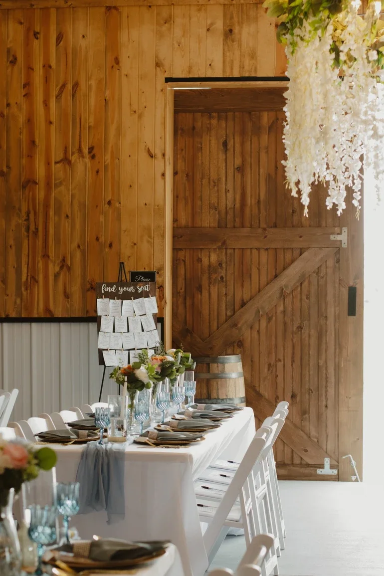 A decorated dining table set for a celebration in a rustic wooden room, with white chairs, floral centerpieces, and a wooden barn door.