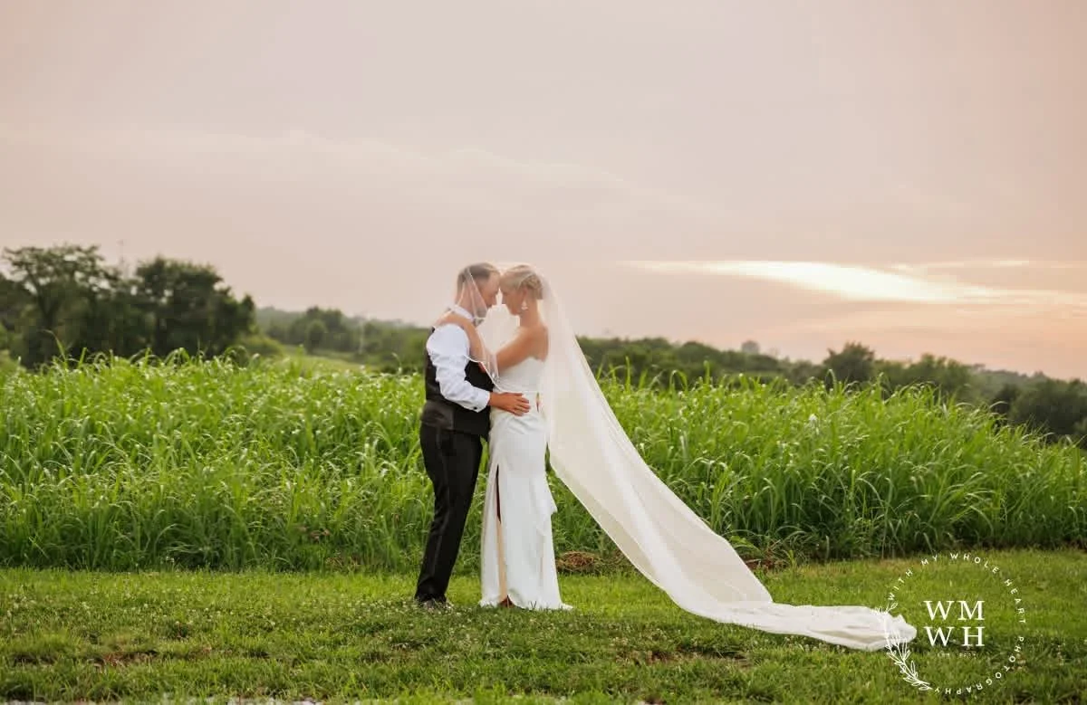 A bride and groom in wedding attire standing close together in a field of green grass at sunset, with the bride wearing a long veil and white dress, and the groom in a black vest and pants.