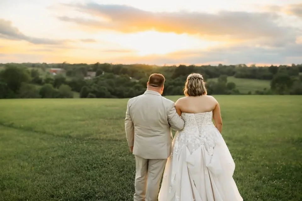 A newlywed couple, with the groom in a light beige suit and the bride in a strapless white wedding gown, walking arm-in-arm across a grassy field at sunset.