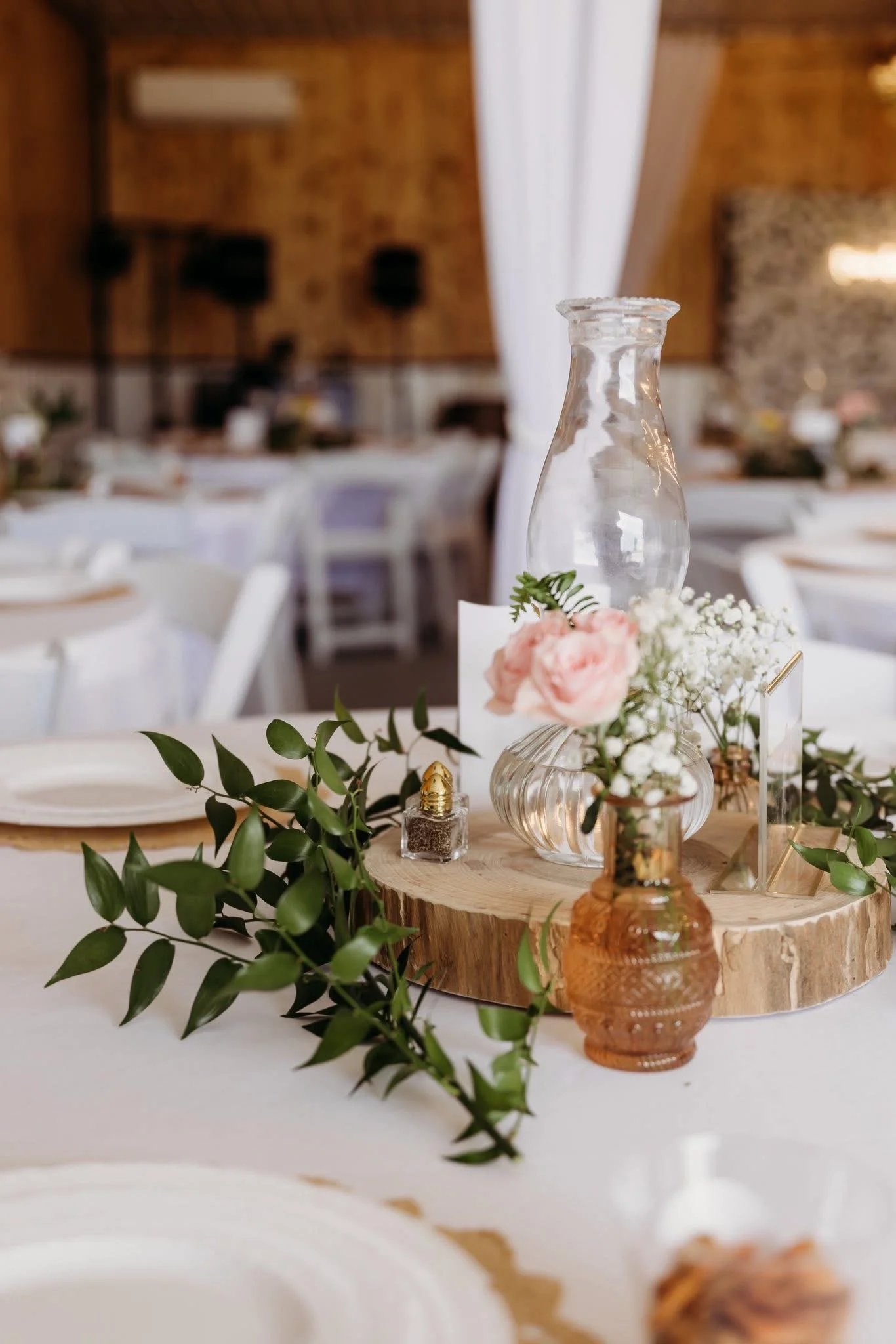 Decorative centerpiece on a table, featuring a clear glass vase with pink and white flowers, surrounded by green foliage and small jars, set on a wooden slab. In the blurred background, a rustic indoor venue with wooden walls and white tables with chairs.