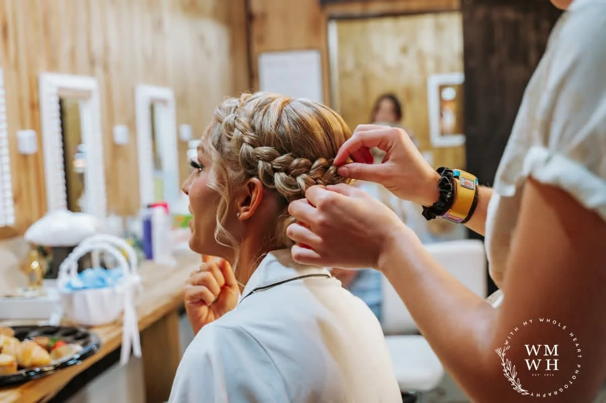A stylist is braiding a woman's blonde hair in a wooden styled room with mirrors and a vanity. The woman is wearing a white robe and smiling as she gets her hair styled.