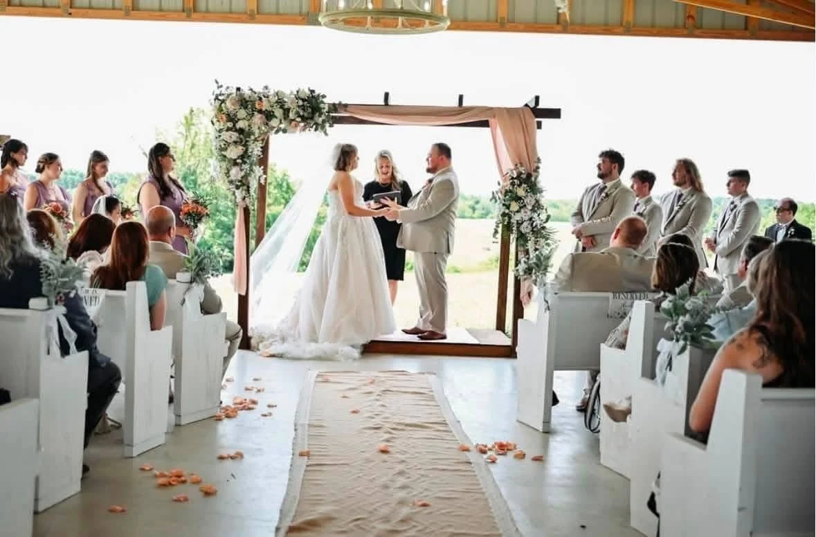 A wedding ceremony taking place indoors with a bride and groom exchanging vows under a decorated arch, surrounded by their wedding party and seated guests.
