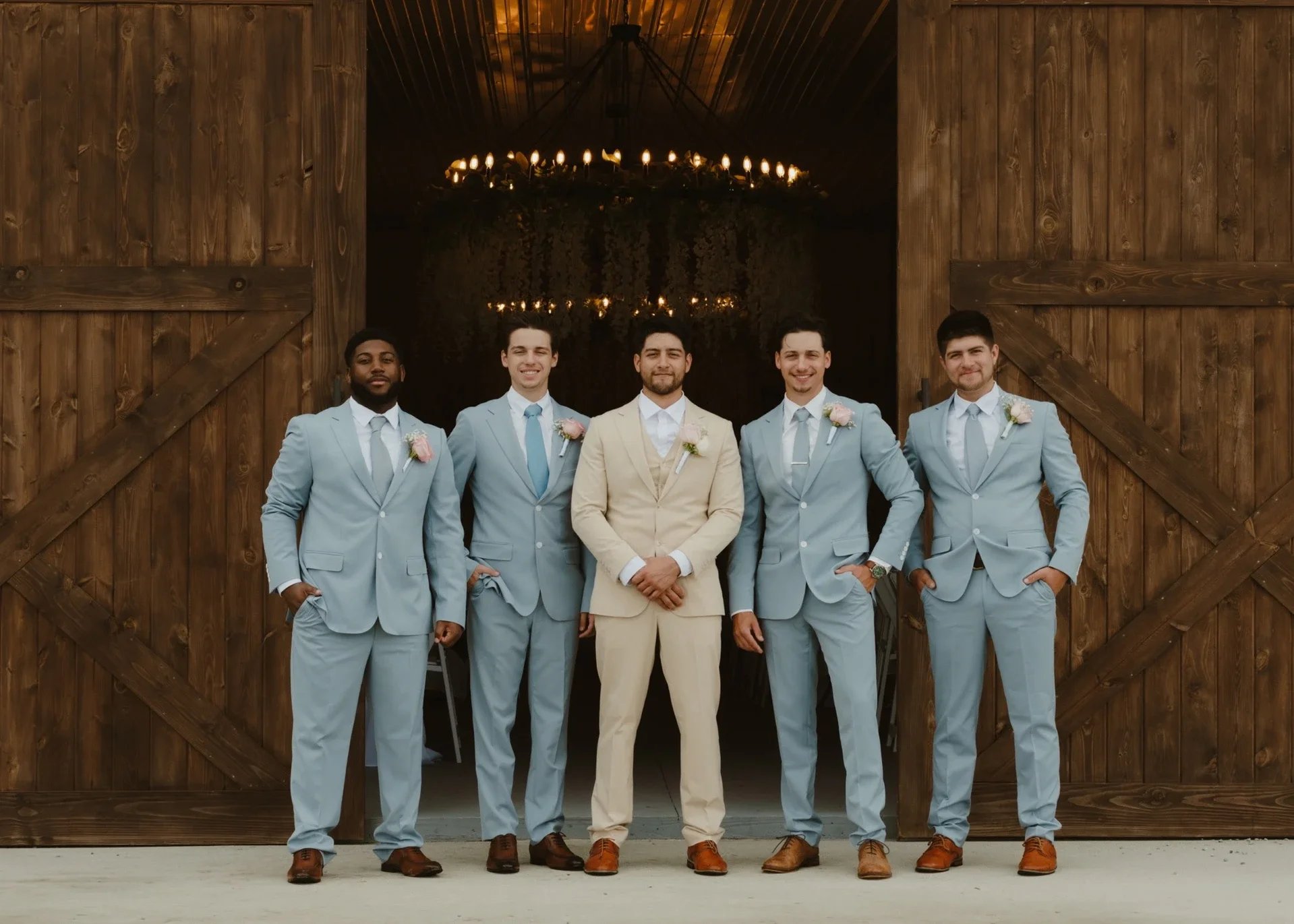 A group of five men dressed in suits, standing in front of a large wooden barn door, with a chandelier hanging inside the barn.