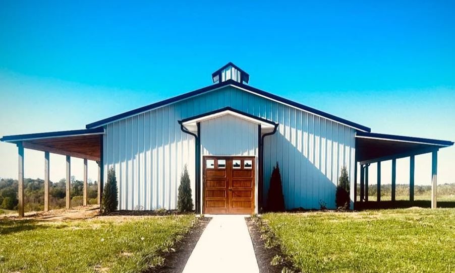 A white barn with a wooden garage door and a small cupola on top, situated on a grassy field with clear blue sky in the background.