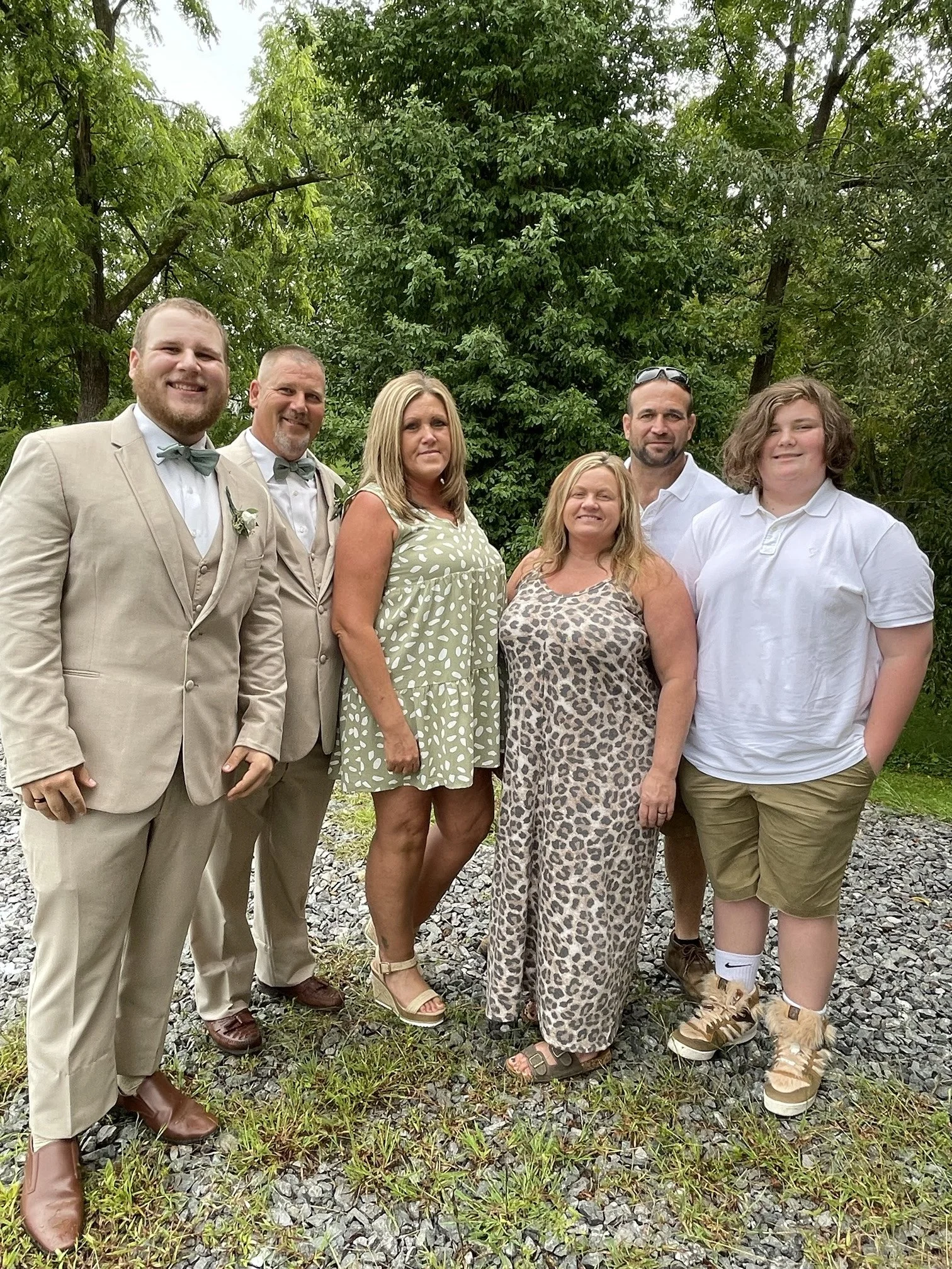 A group of seven people standing outdoors on a gravel area with green trees in the background, dressed in a mix of formal and casual attire.