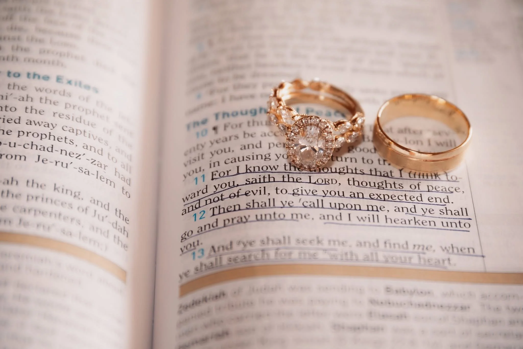 Two gold rings, one with a large central diamond surrounded by smaller diamonds, and the other a plain band, resting on an open Bible with highlighted verses.