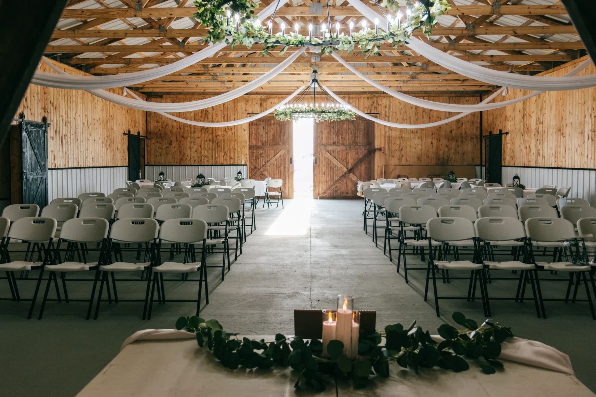 A decorated indoor wedding or event space with wooden walls and ceiling, white drapery, green foliage, a chandelier, and candles on the foreground table, with rows of white chairs arranged facing a large open doorway at the back.