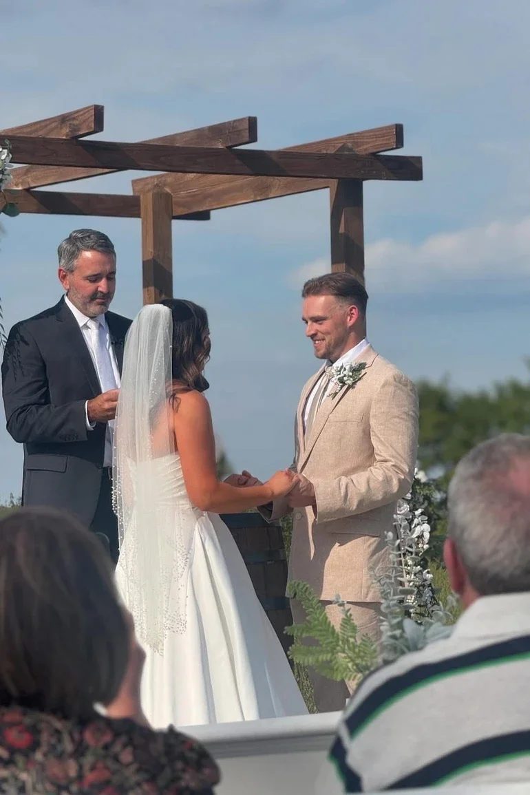 A couple getting married outdoors under a wooden pergola with a man officiating, people in the audience watching.