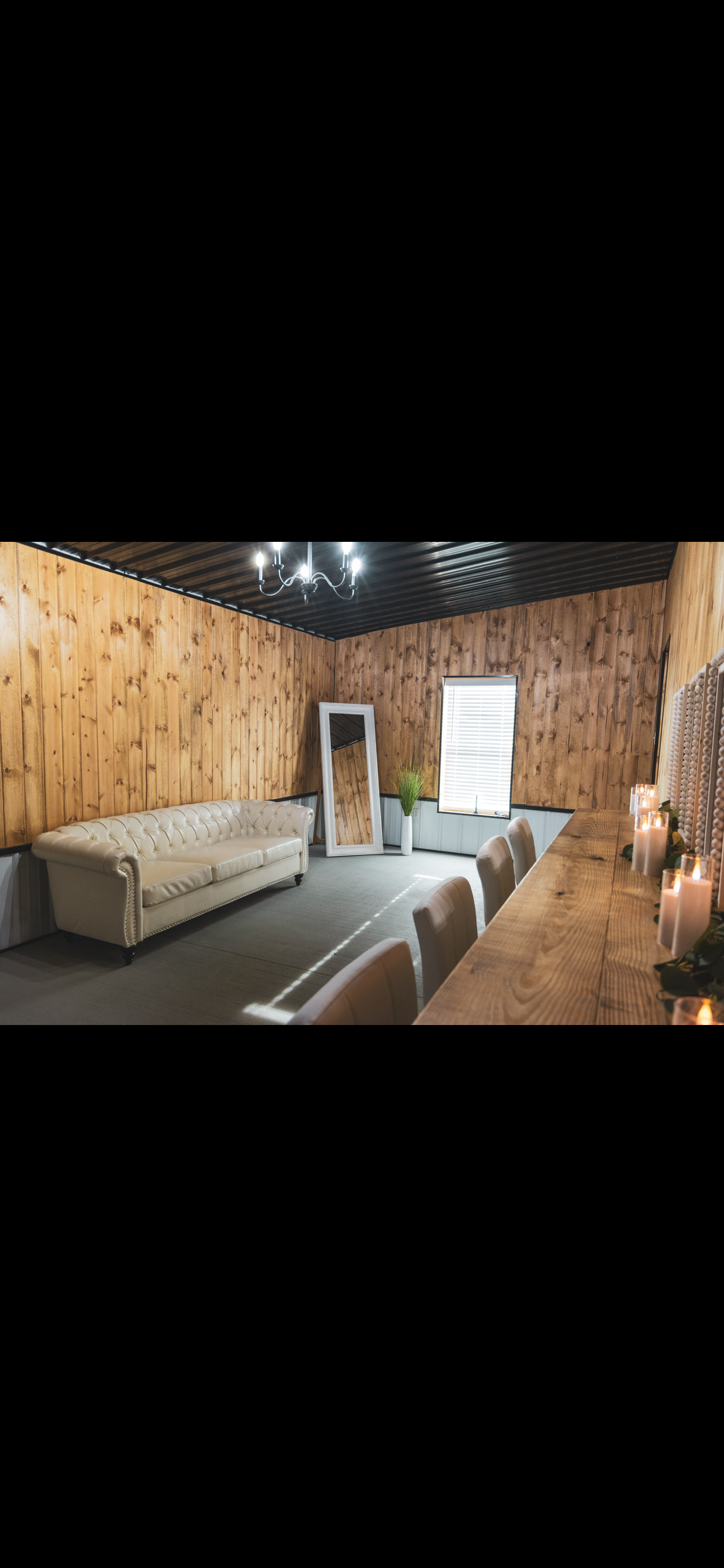 A cozy room with wooden panel walls, a white tufted sofa, a large mirror leaning against the wall, a small potted plant, a window with blinds, a black metal ceiling, a chandelier, and a wooden dining table with candles and chairs.