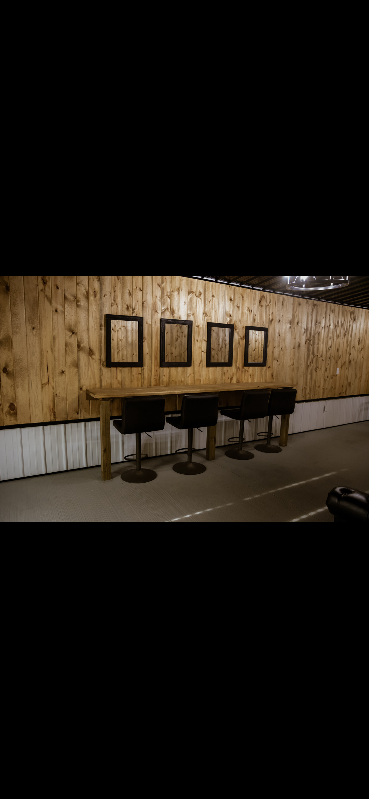 Interior of a room with wooden wall paneling, a long wooden bar-height table with four black bar stools, and four empty black picture frames hanging on the wall.