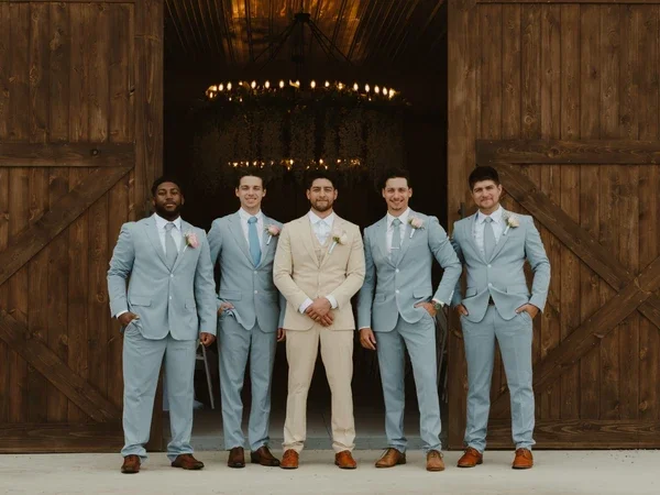 Five men dressed in tuxedos, standing in front of wooden barn doors, with a rustic chandelier hanging in the background.