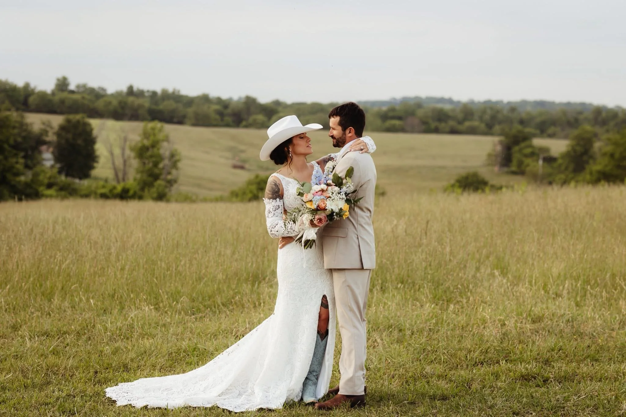 A bride and groom standing closely together in a field, looking into each other's eyes. The bride is holding a bouquet of flowers, wearing a white lace dress and a wide-brimmed hat. The groom is dressed in a light-colored suit. The background features rolling green hills and scattered trees under a partly cloudy sky.