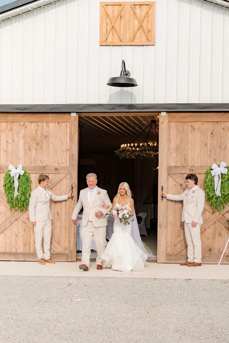 A bride in a white wedding dress holding a bouquet, being escorted out of a barn-style venue by an older man in a cream suit, with two young men in cream suits holding open large wooden barn doors decorated with greenery wreaths with white ribbon bow