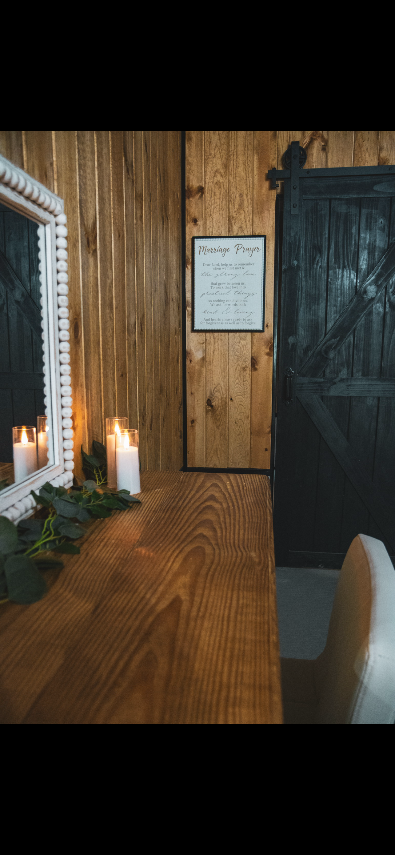 Rustic interior with wooden walls, a black sliding barn door, a white framed mirror, lit candles, and a framed sign reading 'Marriage Prayer' on the wall.