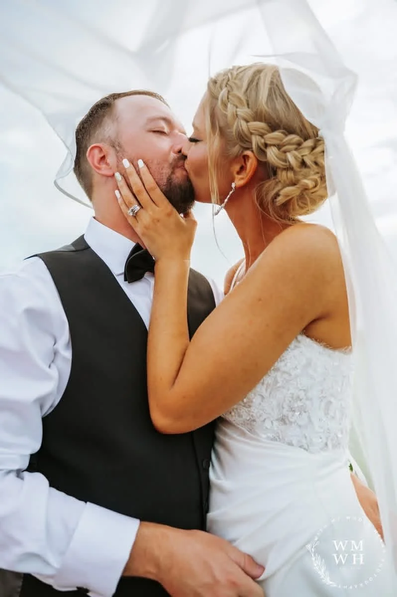 A newlywed couple sharing a kiss outdoors, with the bride wearing a wedding dress and braided hair, and the groom in a tuxedo.