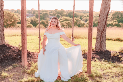 A woman in a white dress sitting on a swing outdoors in a grassy field with trees in the background.