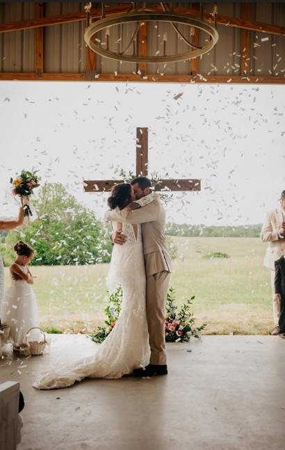 Bride and groom kissing during their wedding ceremony in a rustic barn with a cross in the background, while confetti falls around them.