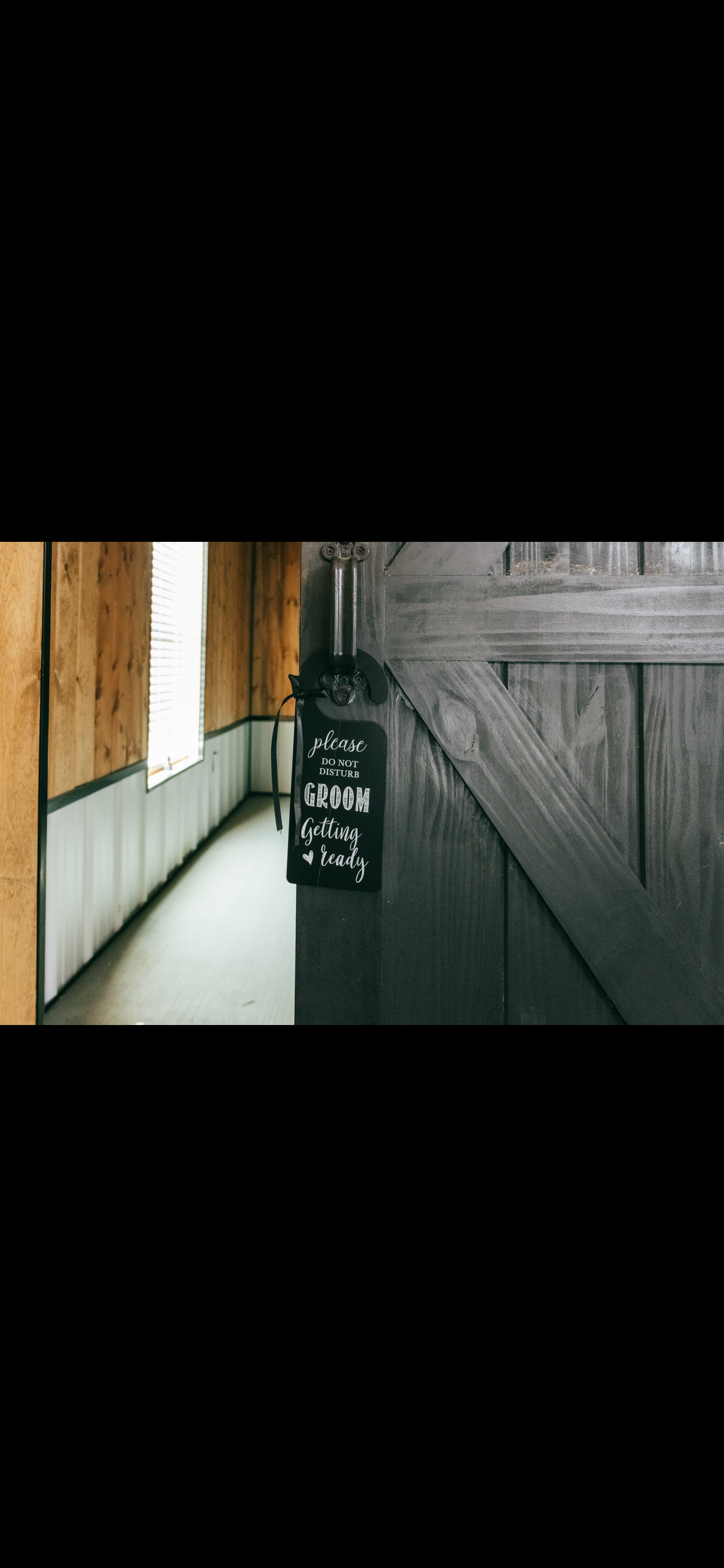 Wooden barn door with a black sign that says "please do not disturb groom getting ready" hanging from the latch. Behind the door, a hallway with wood-paneled walls and a window with white blinds.