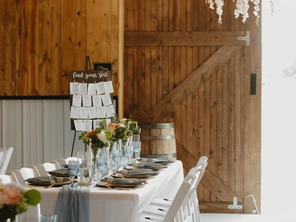Wedding reception setup with a long table, white chairs, floral centerpieces, wine glasses, and place settings, against a wooden wall with a barn door.