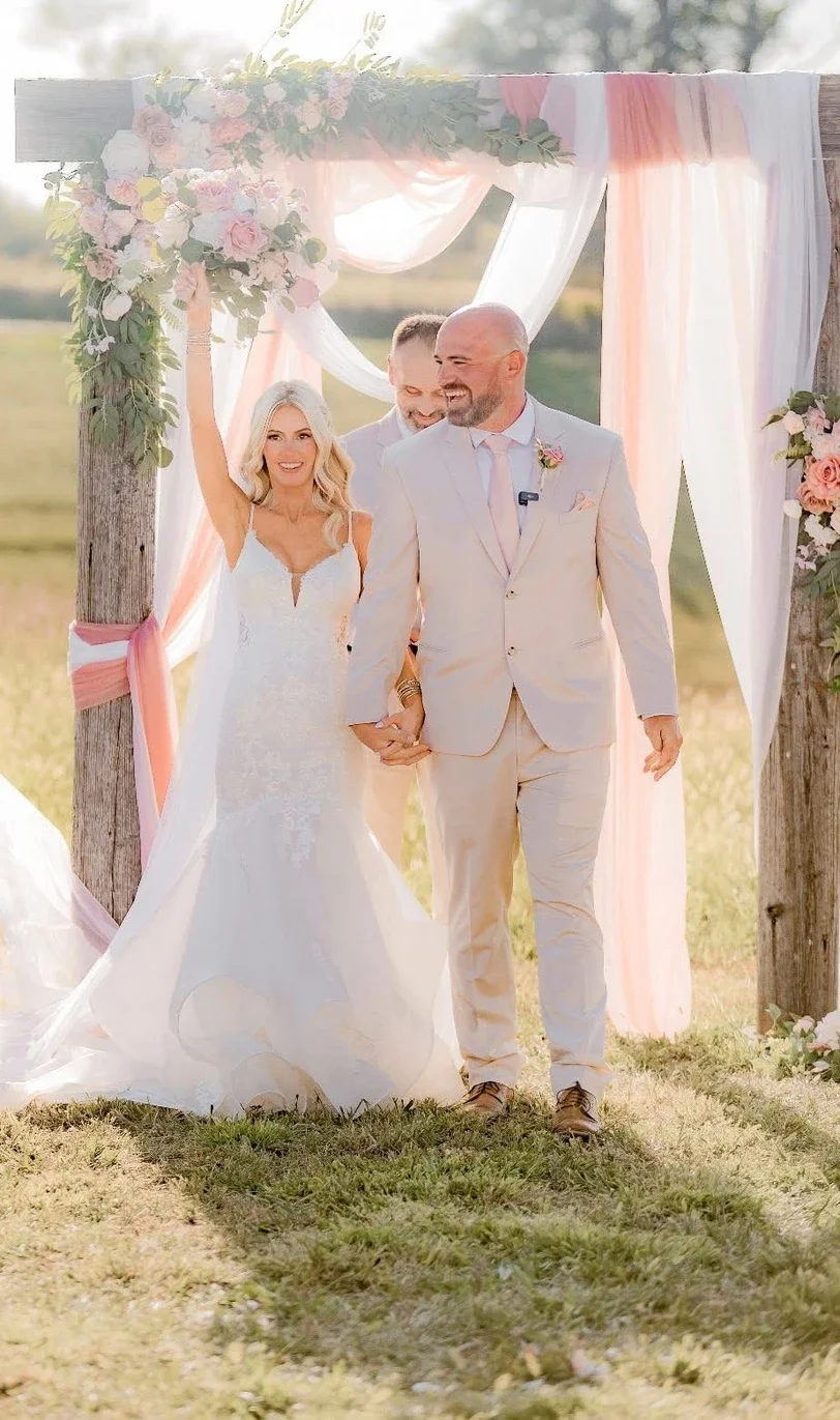 A bride and groom walking hand in hand through a wedding ceremony arch decorated with pink and white flowers, outdoors on grass, with the bride raising her hand in celebration.