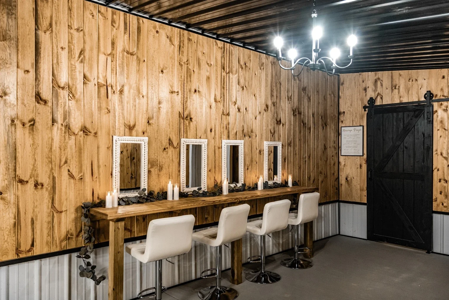 A rustic wooden wall with a long wooden desk, four white chairs, three small mirrors with decorative white frames, candles, and greenery, with a black barn door and a framed sign on the wall. Overhead is a black chandelier with multiple lights.