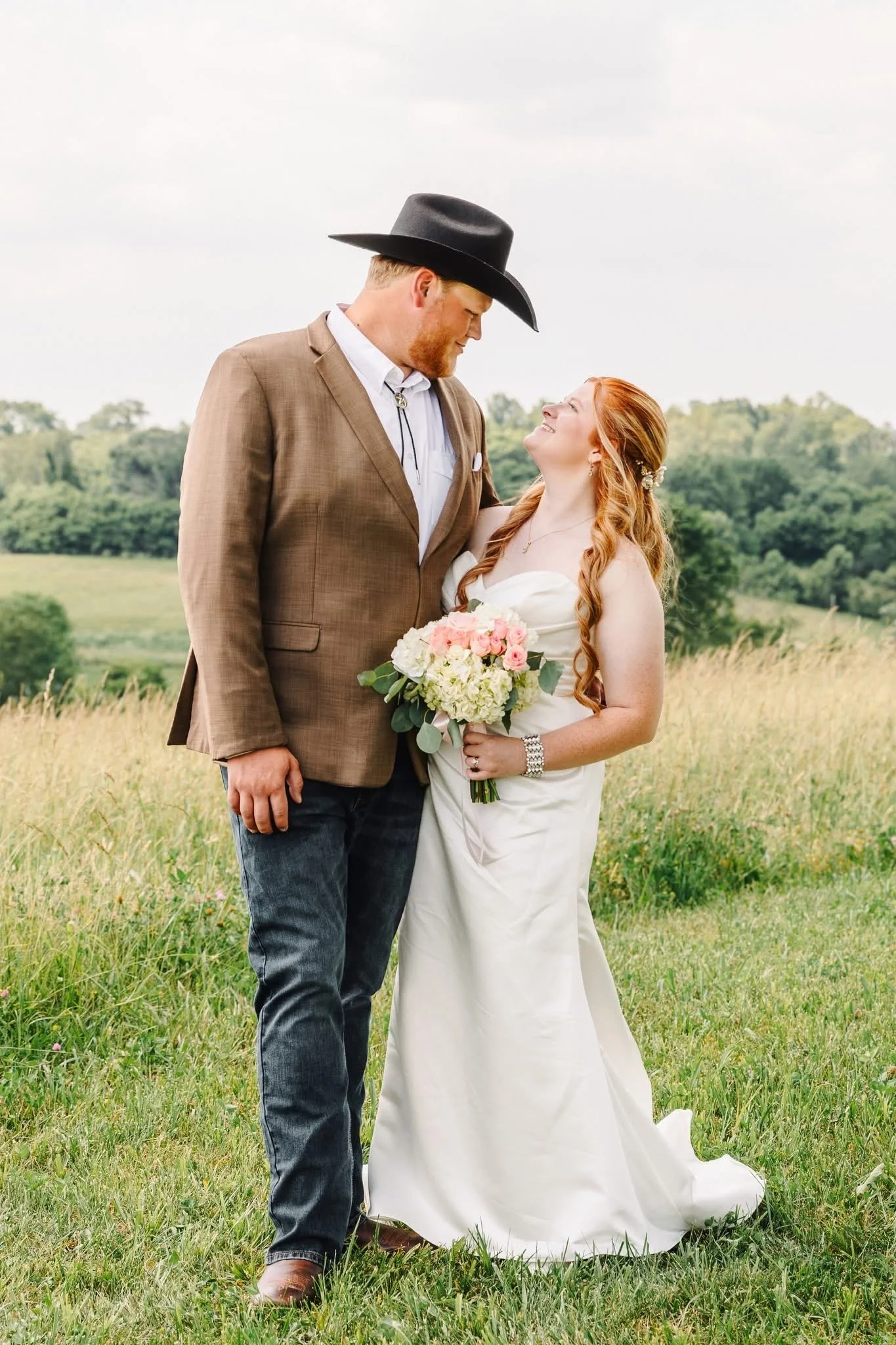 A couple dressed in wedding attire outdoors in a grassy field with trees in the background, smiling and looking into each other's eyes, holding a bouquet of flowers.