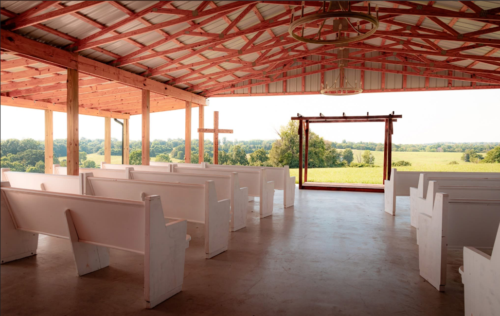 An open-air chapel with white wooden pews, a wooden altar frame, and a cross on a scenic green landscape background.