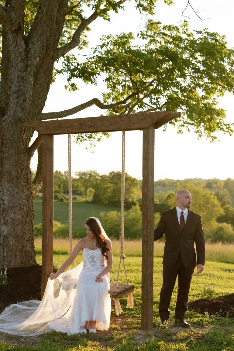Bride in a white wedding dress sitting on a wooden swing hanging from a large tree, groom in a suit standing nearby outdoors in a grassy field during sunset.