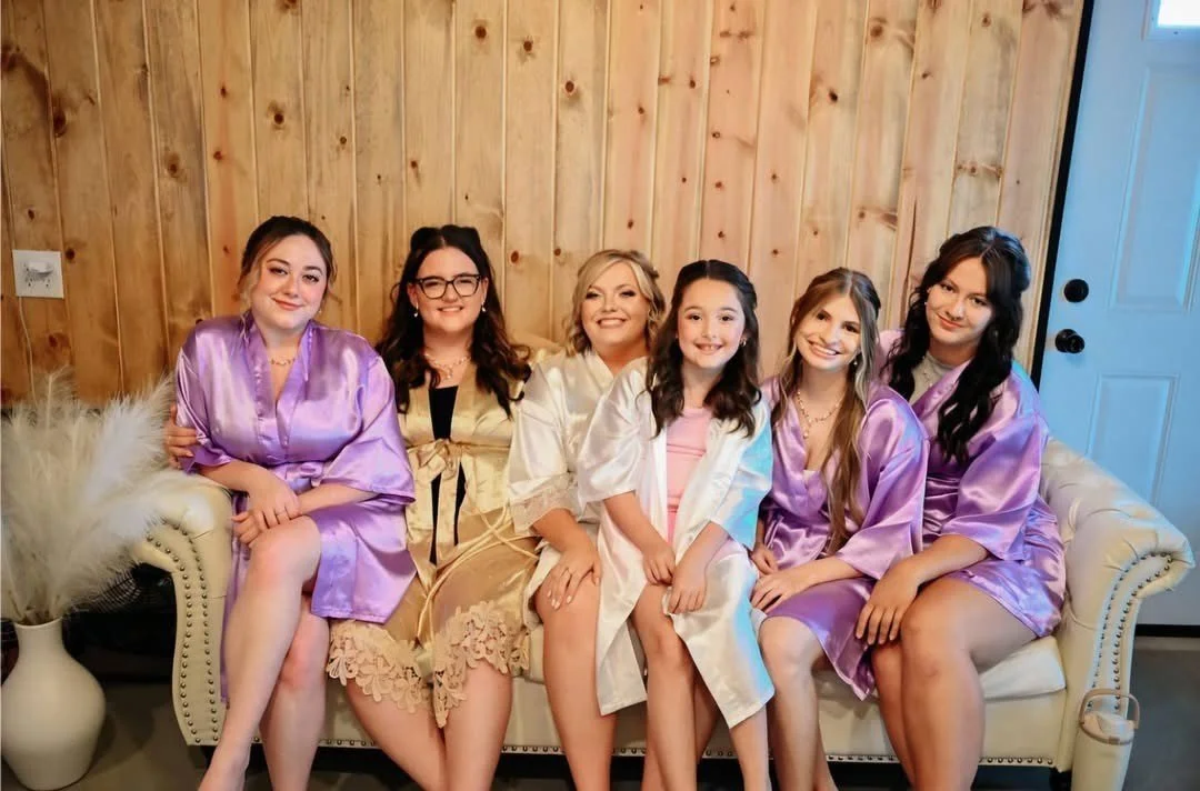 Six women sitting on a beige couch, dressed in pastel-colored robes, in a room with wooden walls.