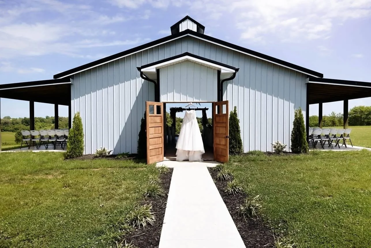 A white building with black roof accents, open wooden double doors, and a wedding dress hanging inside, with a pathway leading to the entrance surrounded by small plants and a grassy yard under a partly cloudy sky.